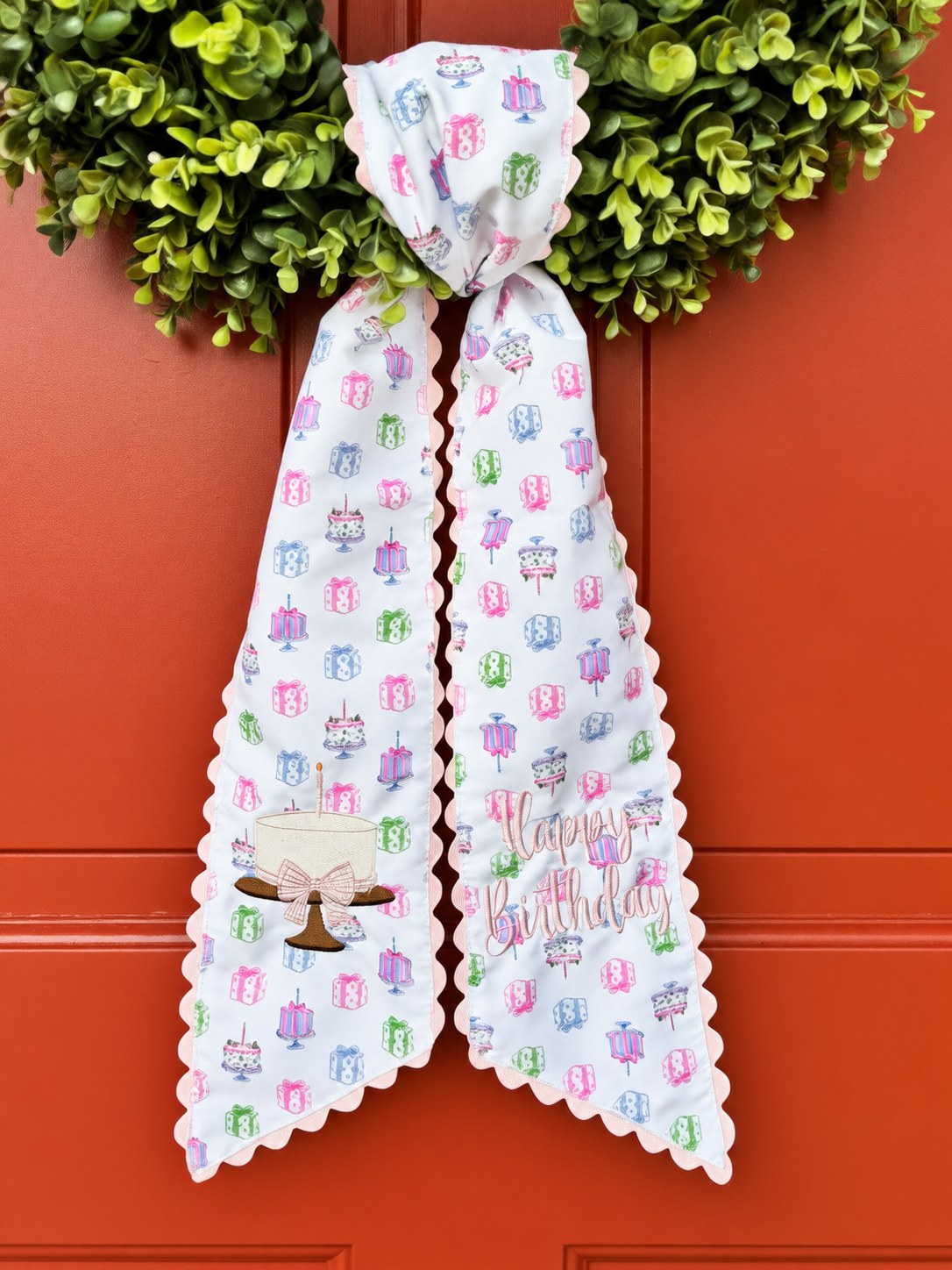 White wreath sash printed with birthday cakes and gifts, embroidered with a cake and bow and Happy Birthday, tied to a boxwood wreath on a red door