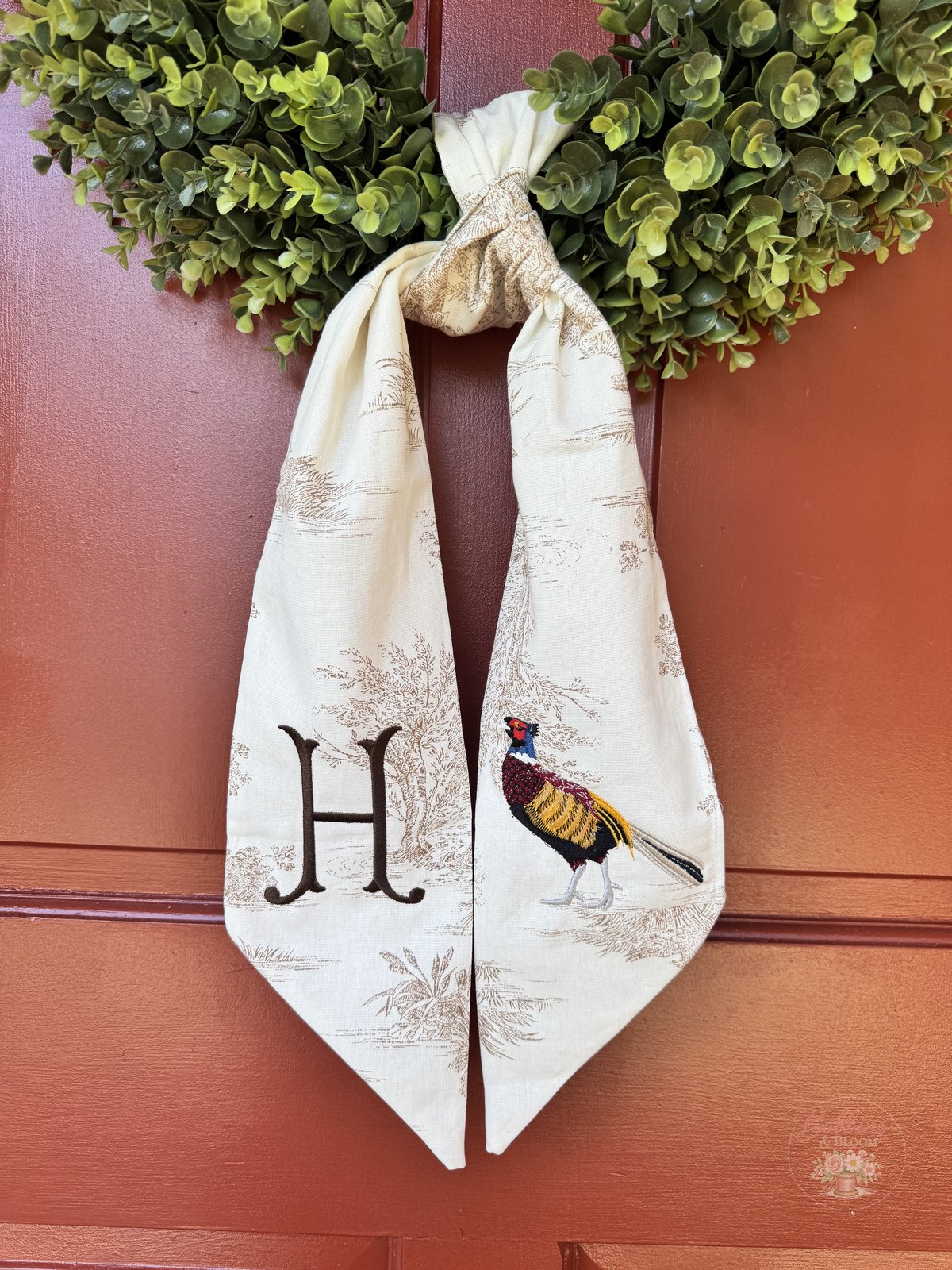Cream toile wreath sash with an embroidered pheasant and H monogram, tied to a boxwood wreath